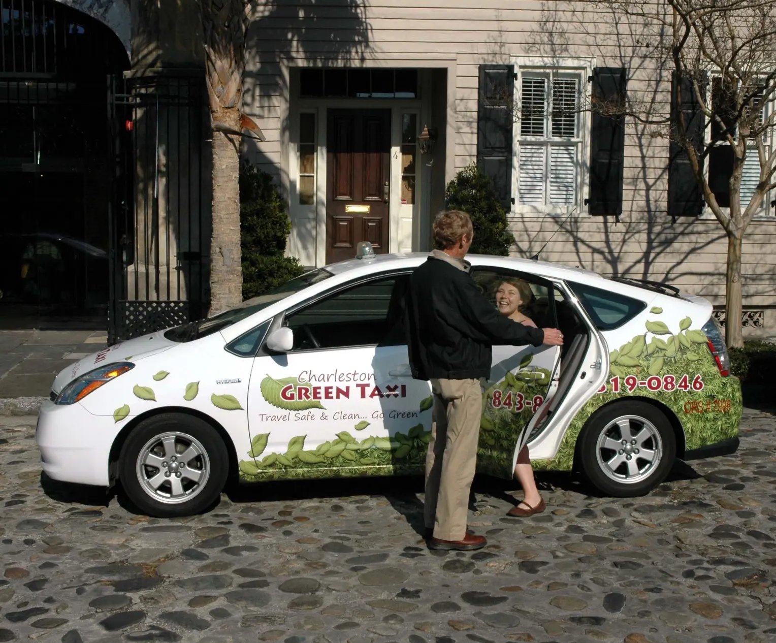 A Charleston Green Taxi driver holding the door for a passenger on a cobblestone downtown street