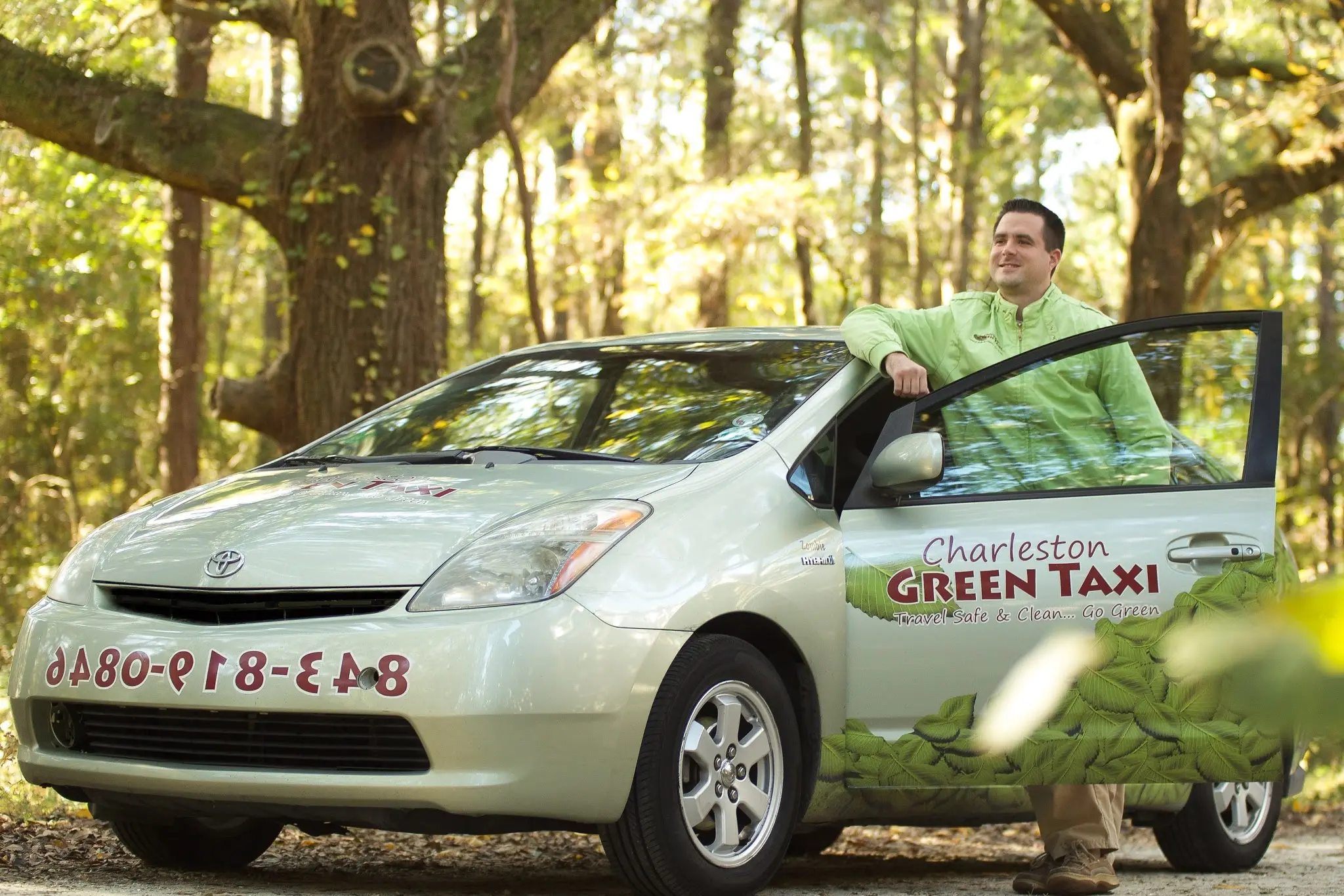 A Charleston Green Taxi driver standing with an early wrapped Prius on a Lowcountry dirt road under the oaks